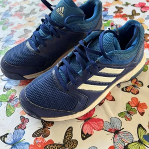 Blå Adidas sneakers med vita ränder - Snygga blå sneakers från Adidas med klassiska vita ränder på sidan. Ovandelen är i mesh och syntet för bra andningsförmåga och komfort. Skorna har snörning och en platt sula, perfekt för dig som gillar sportig stil och vill ha bekväma skor till vardagen.