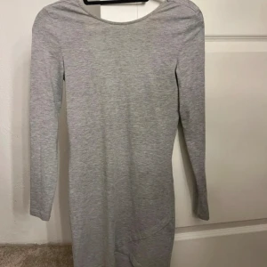 Grå långärmad klänning i bomull - En enkel och stilren grå klänning med lång ärm och rund halsringning framtill samt djupare urringning i ryggen. Klänningen är figurnära och tillverkad i mjuk bomull, perfekt för en clean och avslappnad look.