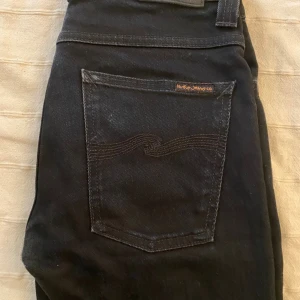 Svarta jeans från Nudie Jeans Co - Säljer ett par svarta jeans från Nudie Jeans Co med klassisk femficksdesign och snygga svarta sömmar på bakfickan. Jeansen har en stilren look med svarta detaljer och patch bak i midjan. Perfekta för dig som gillar minimalistisk stil.