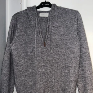 Grå stickad hoodie från Pier One - Mysig grå stickad hoodie från Pier One med huva, snörning och half zip-dragkedja framtill. Tröjan har lång ärm och ribbade muddar vid ärmslut och nederkant. Perfekt för kyliga dagar och enkel att matcha med jeans eller joggers.