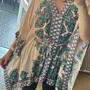 Mönstrad kaftan i satin viskos - Superfin luftig kaftan i ljusbeige med grönt och svart paisleymönster. Kaftanen har v-ringning, vida ärmar och knytskärp i midjan. Viskos satin. XS/S i storlek men passar även M. Skärpet går att ta av. 