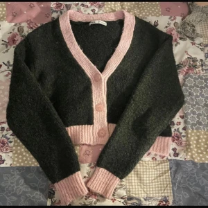 Mörkgrå och rosa kofta från Zara - Supermysig croppad kofta från Zara i mörkgrått med breda ljusrosa ribbade detaljer vid knappar, ärmslut och nederkant. Koftan har v-ringning, stora knappar framtill och är stickad i ett mjukt material. Perfekt för lager-på-lager under kyliga dagar.