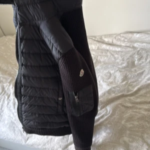 Svart Moncler pufferjacka M - Säljer en svart pufferjacka från Moncler i storlek M. Jackan har stickade ärmar och quiltad kropp, med dragkedja och knappdetaljer. Klassisk Moncler-logga på ärmen och en liten ficka med dragkedja på ena ärmen. Perfekt för kyliga dagar.