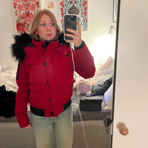 Röd pufferjacka med svart fuskpäls - Röd pufferjacka från Hollister med svart fuskpäls på huvan och svarta detaljer. Jackan har stora fickor framtill, ribbade muddar och dragkedja med knappar. Perfekt för kalla dagar för alla mina 2000’s babes! 