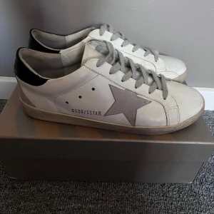 Golden Goose Superstar sneakers vit/grå - Golden Goose Superstar sneakers i vitt skinn med grå mockastjärna på sidan, grå snörning och svart häl. Klassisk låg modell med platt sula och rund tå. Snygg och avslappnad stil som passar till det mesta. Skokartong medföljer. står 40 som skostorlek men passar ÄVEN upp till 42 pga de är ganska stora i storlek.