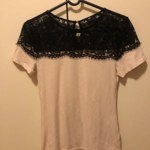 Vit topp med svart spets från H&M - Snygg vit topp från H&M med korta ärmar och svart spets upptill. Toppen har rund halsringning och en elegant spetsdetalj över axlar och bröst. Perfekt för dig som gillar kontraster och stilrena detaljer.