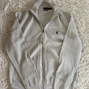 Ljusgrå kofta från Polo Ralph Lauren - Ljusgrå kofta från Polo Ralph Lauren i mjuk pima bomull. Tröjan har hel dragkedja, ribbade muddar och diskret logga broderad på bröstet. Perfekt för lager-på-lager och har en clean, stilren look.