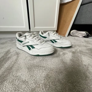 Vita och gröna Reebok sneakers - Säljer ett par klassiska Reebok sneakers i vitt med gröna detaljer och logga på sidan och hälen. Skorna har snörning, perforerad tå och grön sula. Perfekta för dig som gillar retrostil och vill ha ett par stilrena och bekväma sneakers i skinn.