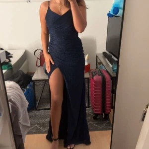 Marinblå glittrig långklänning med slits - Säljer en marinblå långklänning med glittrigt tyg och en hög slits på ena benet. Klänningen har smala axelband och figurnära passform som framhäver siluetten. Perfekt för bal eller festkvällar när du vill sticka ut.