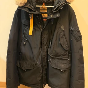 Svart Parajumpers vinterjacka XL - Svart vinterjacka från Parajumpers med pälsfodrad huva, flera fickor och ikonisk gul rem vid kragen. Jackan har dragkedja, knappar och detaljer i svart och gult. Slim fit-modell, perfekt för kalla dagar. Snygg och funktionell design.