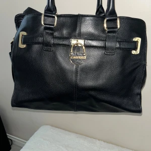 Svart handväska från Calvin Klein - Stilren svart handväska från Calvin Klein i äkta skinn med guldfärgade metalldetaljer. Väskan har dubbla handtag och ett dekorativt spänne med logotyp framtill. Perfekt storlek för att få plats med allt du behöver och passar till många olika looks.