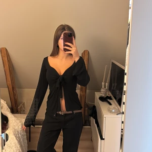 Svart omlottopp med knyt från Design by Si - Svart långärmad omlottopp från Design by Si med djup v-ringning och knyt framtill. Toppens material känns mjukt och stretchigt, perfekt för en trendig look. Passformen är normal och toppen har en croppad känsla med öppen front.