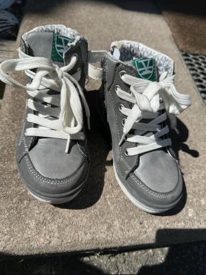 Grå höga sneakers med vita snören - Snygga grå sneakers med högt skaft och vita snören. Skorna har vita sömmar och en grön logga på plösen. Insidan har ett diskret mönster i vitt och grått. Perfekta för dig som gillar en stilren och sportig look.