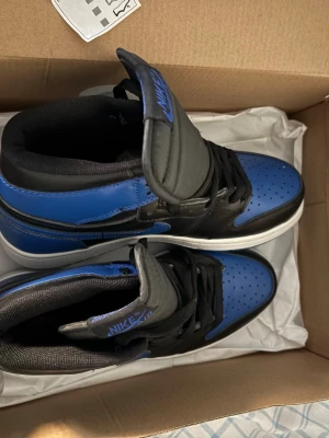 Nike Air Jordan 1 High svart/blå - Säljer ett par Nike Air Jordan 1 High sneakers i svart och blått med vit sula. Skorna har klassisk snörning, perforerad tå och ikonisk Air Jordan-logga på sidan. Materialet är skinn och syntet, och färgkombon ger en riktigt snygg vibe. Perfekt för dig som gillar streetwear och vill sticka ut.