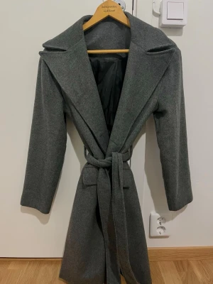 Grå kappa med bälte och bred krage - Snygg grå kappa med bred krage och knytbälte i midjan. Långärmad och rak modell som ger en clean och stilren look. Perfekt för kyligare dagar och passar till det mesta. Fodrad insida för extra komfort.