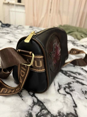 Svart axelväska med lotusmönster - Cool svart axelväska från Qianxidaishu i skinn med ett detaljerat lotusblommotiv i rosa och lila på framsidan. Väskan har bred, justerbar axelrem i brunt med logotyp, guldfärgad dragkedja och flera fack inuti. Perfekt för dig som gillar unika detaljer och snygg design.