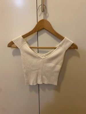 Vit ribbad croppad topp - Säljer en vit ribbad croppad topp med breda axelband och v-ringning. Går bra att ha som linne med v-ringning eller som off-shoulder. Strl XS
