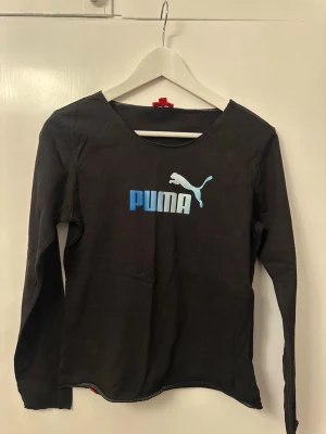 Svart långärmad topp från Puma - Svart långärmad topp från Puma med stor logga i blått och vitt framtill. Tillverkad i mjuk bomull med rund halsringning. Perfekt för en sportig och avslappnad stil.