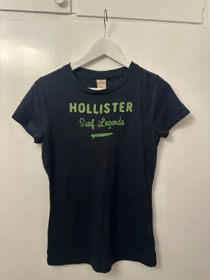 Svart Hollister t-shirt med brodyr - Snygg svart t-shirt från Hollister med grön broderad text 'Surf Legends' på bröstet. Klassisk rund hals och korta ärmar. Tillverkad i mjuk bomull som känns skön mot huden. Perfekt för dig som gillar en enkel men stilren look.