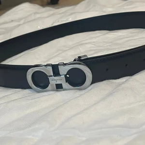 Svart Ferragamo bälte med silverdetalj - Snyggt svart bälte från Ferragamo med ikoniskt, rundat spänne i silverfärgad metall. Bältet är gjort i slätt skinn och har en stilren, klassisk design som passar till många outfits. Perfekt accessoar för att lyfta din look.