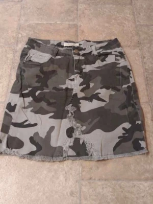 Camouflage kortkjol med fransad kant - Säljer en cool kortkjol i camouflage-mönster med fransad kant och slitningar framtill. Kjolen har fickor och bälteshällor, perfekt för dig som gillar streetstyle och vill sticka ut lite extra.