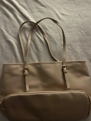 Beige axelväska med justerbara remmar - Snygg beige axelväska med stilrena justerbara remmar och guldfärgade spännen. Väskan har en enkel, clean design och ett stort fack framtill. Perfekt för dig som vill ha en neutral och praktisk väska till vardagen.