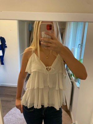 Vit volangtopp med knyt - Säljer en superfin vit linnetopp med tunna justerbara axelband och flera lager volanger. Toppen har även knytning framtill och är lätt och luftig. Kom privat för fler frågor eller bilder!💕