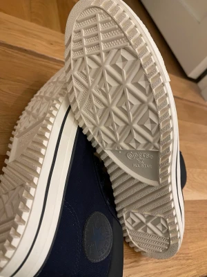 Mörkblå Converse All Star high-tops - Säljer ett par mörkblå Converse All Star high-tops med vit sula och tå, samt svarta detaljer. Skorna har snörning, vadderad kant och slitstark canvas. Klassisk Converse-logga på sidan och grovt mönstrad sula för extra grepp. Perfekta för dig som gillar streetstyle.