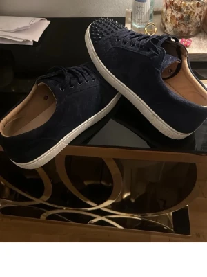 Mörkblå loubs sneakers med nitar - Säljer ett par mörkblå sneakers med coola svarta nitar på tån. Skorna har snörning och en vit platt sula. Utsidan är i mocka och insidan är ljusbeige. Perfekt för dig som vill sticka ut med din stil.