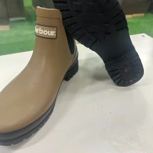 Beige Barbour Chelsea boots - Snygga beige Chelsea boots från Barbour med svart grov sula och elastiska paneler på sidorna. Skorna har en rund tå och är tillverkade i syntetmaterial som tål regn. Perfekt för dig som vill ha en stilren look med tydlig logga framtill.