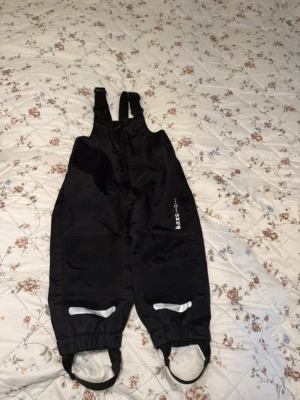 Svarta täckbyxor från Kaxs - Svarta täckbyxor från Kaxs med hängslen och dragkedja framtill. Byxorna har resår vid benslut och praktiska fothällor. Perfekta för kalla dagar och utomhusaktiviteter. Reflekterande detaljer på benen för extra synlighet.