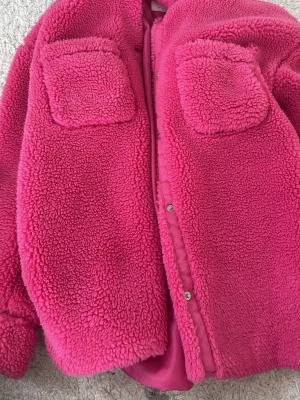 Rosa teddyjacka från H&M - Säljer en fluffig och cool rosa teddyjacka från H&M med två stora fickor framtill och tryckknappar. Jackan har en oversized look och är gjord i mjuk fleece som håller dig varm och skön. Perfekt för dig som gillar färg och vill sticka ut.