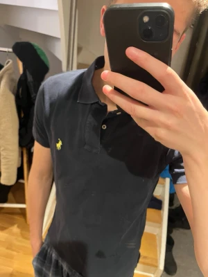 Svart pikétröja från Ralph Lauren - Klassisk svart pikétröja från Ralph Lauren med gul broderad logga på bröstet. Tröjan har krage, korta ärmar och knäppning med två knappar framtill. Tillverkad i bomull och har en normal passform.