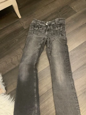 Grå jeans med fickdetaljer - Snygga grå jeans med coola fickor framtill som har lock och knappdetaljer. Jeansen har en rak passform och klassisk femficksdesign. Materialet är jeans och färgen är mörkgrå med lite slitningar för en schysst look.