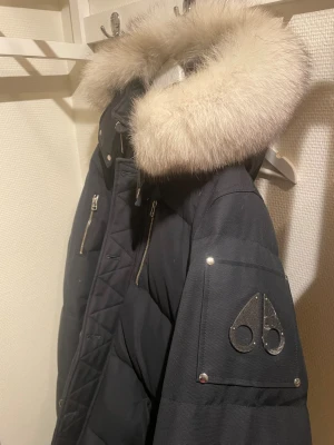 Svart Moose Knuckles dunjacka med päls - Säljer en svart dunjacka från Moose Knuckles med fluffig ljus päls på huvan. Jackan har flera dragkedjor, quiltade detaljer och en snygg logga på ärmen. Perfekt för kalla dagar och har en riktigt schysst look med sina silvriga metalldetaljer.