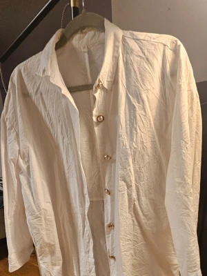 Vit skjorta med pärlknappar, strl M - Säljer en vit skjorta i storlek M med unika pärlknappar med gulddetaljer. Skjortan har lång ärm och klassisk krage, tillverkad i ett lätt och skönt bomullsmaterial. Perfekt för dig som gillar stilrena plagg med en twist på detaljerna.