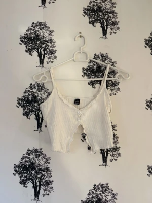 Vit ribbad croppad linnetopp Shein - Säljer en vit croppad linnetopp från Shein i storlek XS. Toppen har smala axelband, ribbad struktur och knappar framtill. Den har en vågig, rå kant nertill som ger en trendig look. Perfekt till höga jeans eller kjol.