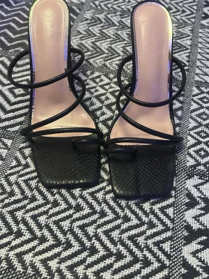 Svarta sandaler med hög klack - Säljer ett par svarta sandaler med smala remmar och fyrkantig tå. Skorna har hög klack och en snygg struktur i ormskinnsmönster. Perfekta för dig som vill ha en trendig och elegant look.