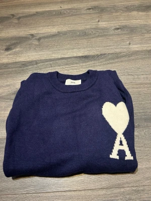 Mörkblå stickad tröja från Ami - Mörkblå stickad tröja från Ami med vit hjärtlogga och bokstaven A på bröstet. Tröjan har rund halsringning, långa ärmar och ribbade muddar. Perfekt för dig som gillar stilrena detaljer och vill ha något som sticker ut lite.