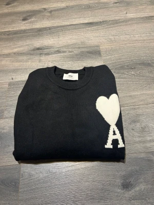 Svart stickad tröja från Ami - Svart stickad tröja från Ami med rund hals och lång ärm. På bröstet finns ett stort vitt hjärta och bokstaven A som ger en unik och trendig look. Tröjan har ribbade muddar vid hals, ärmslut och nederkant. Perfekt för dig som gillar stilrena detaljer.