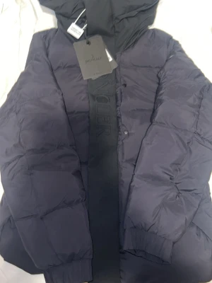Svart Moncler pufferjacka med huva - Säljer en svart pufferjacka från Moncler med stor huva och broderad logga på ärmen. Jackan har en bred frontpanel med Moncler-text, ribbade muddar och knappar framtill. Perfekt för kalla dagar och riktigt snygg streetstil.