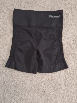 Svarta träningsshorts från Hummel - Säljer ett par svarta, högmidjade träningsshorts från Hummel. De är tillverkade i ett stretchigt syntetmaterial med ribbade paneler på sidorna för extra komfort. Perfekta för gymmet eller löpning, med diskret logga framtill.
