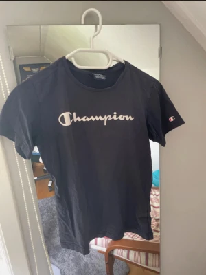 Mörkblå Champion t-shirt - Mörkblå t-shirt från Champion med klassisk logga i vitt på bröstet och liten logga på ärmen. Modellen är kortärmad och har en rund halsringning. Materialet är mjuk bomull som känns skön mot huden. Perfekt för en chill och sportig stil.