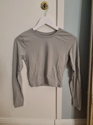 Grå croppad långärmad topp 157 - Säljer en stilren grå croppad topp från 157 i storlek S. Toppen har lång ärm och är tillverkad i mjuk bomull med lite elastan för extra stretch. Enkel design med rund hals och tight passform, perfekt att matcha med jeans eller kjol.