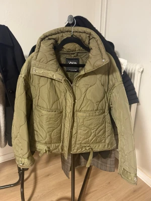 Quiltad grön jacka från Zara - Säljer en quiltad jacka från Zara i en snygg olivgrön färg. Jackan har hög krage, två stora fickor framtill och dragkedja. Den är croppad med snörning vid midjan och ärmarna, perfekt för att ge en cool vibe till din outfit. Använd två gånger efter att ha köpt på Vinted och ser helt ny ut