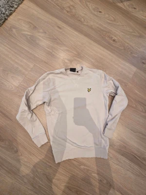 Beige sweatshirt från Lyle & Scott - Säljer en beige sweatshirt från Lyle & Scott med deras klassiska gula fågel-logga broderad på bröstet. Tröjan har rund hals, långa ärmar och ribbade muddar vid ärmslut och nederkant. Perfekt för chill dagar och enkel att matcha.