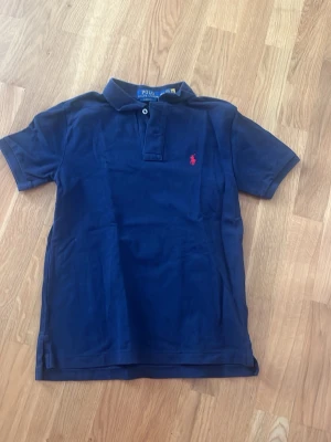 Marinblå piké från Polo Ralph Lauren - Säljer en klassisk marinblå pikétröja från Polo Ralph Lauren med röd broderad logga på bröstet. Tröjan har korta ärmar, krage och två knappar vid halsen. Classic fit-modell i mjuk bomull som är perfekt för en chill och stilren look.