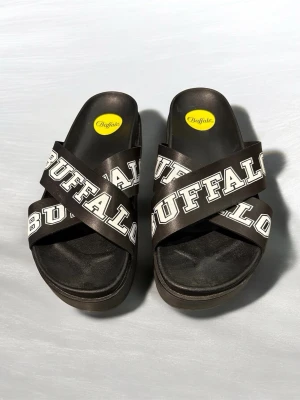 Buffalo svarta platåsandaler - Buffalo sandaler med breda svarta remmar i syntetmaterial och vit logotyptext. Platåsula för extra höjd och chunky vibe. Perfekta för stranden eller cityhäng. Klassisk rund tå och öppen design för skön sommarkänsla.