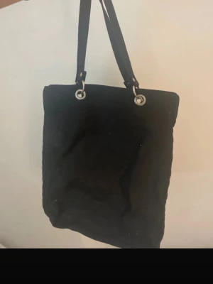 Svart axelväska i bomull - Säljer en svart axelväska i bomull med två rejäla handtag och silverfärgade metallringar. Väskan har en enkel, stilren design och en praktisk dragkedja på baksidan. Perfekt storlek för att få plats med allt du behöver till skolan eller stan.