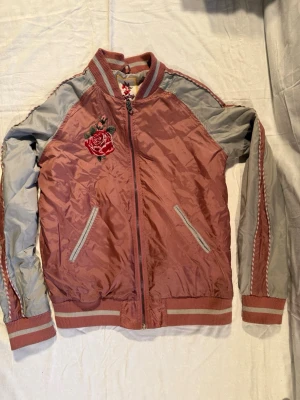 Rosa bomberjacka från Replay - Snygg bomberjacka i rosa och grått från Replay med broderade blommor på bröstet och ryggen. Jackan har ribbade muddar, dragkedja framtill och två fickor. Materialet är glansigt och känns lätt på kroppen. Perfekt för dig som vill sticka ut lite extra.